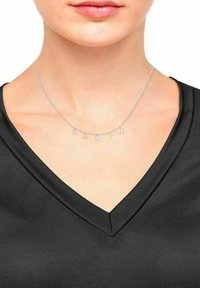 Silberne Halskette mit Buchstaben, die "ERDE" auf einer feinen Kette buchstabieren, getragen zu einem schwarzen V-Ausschnitt-Oberteil. Glatte Metalloberfläche, minimalistische Gestaltung.