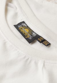 Hvid bomulds T-shirt med ribbet halsudskæring, med et sort mærke broderet med en sol og palmer i guld samt hvid tekst.