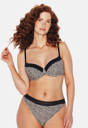 Femme aux cheveux roux bouclés portant un soutien-gorge et une culotte assortis à imprimé léopard noir et blanc, posant avec une main touchant ses cheveux.