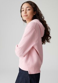 Felpa rosa con cappuccio dalla vestibilità comoda, polsini e orlo a coste. Tessuto morbido con dettagli di cuciture visibili. Indossata sopra jeans scuri.