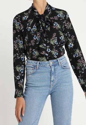 Persoon die een zwarte blouse met lange mouwen draagt met een blauw en roze bloemenprint en lichtblauwe high-waisted jeans met knopen aan de voorkant en zakken.