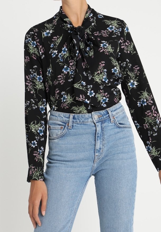 Personne portant une blouse noire à manches longues avec un imprimé floral bleu et rose et un jean taille haute bleu clair avec bouton et poches à l'avant.