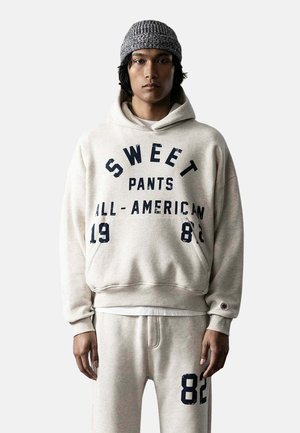 Persona joven con gorro de punto gris, sudadera con capucha beige clara con el texto "Sweet Pants All-American 1982" y pantalones deportivos a juego con el número 82.