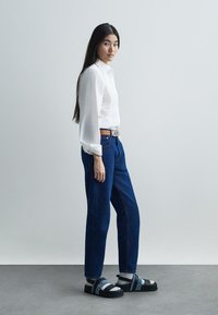 Fehér gombos ing, kék magas derekú farmernadrág, fekete szandál kék pántokkal, világosszürke háttér előtt állva.