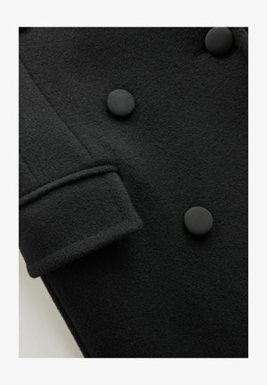 Szczegóły czarnego wełnianego płaszcza z podwójnym zapięciem, teksturowaną tkaniną, okrągłymi czarnymi guzikami i złożonym mankietem.
