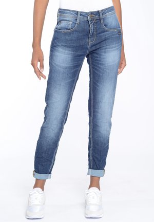 Nainen, jolla on ylös käännetyt siniset farkut ja valkoiset lenkkarit, seisoo yksinkertaisen valkoisen taustan edessä.