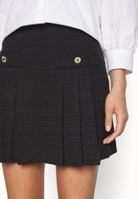 Mini-jupe plissée noire avec deux boutons dorés sur les poches avant, portée avec une chemise blanche à manches longues et manches retroussées.