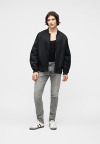 Νέος άνδρας όρθιος, φορόντας μαύρο μπουφάν τύπου bomber, μαύρο αμάνικο μπλουζάκι, ανοιχτό γκρι τζιν και λευκά αθλητικά παπούτσια με μαύρες ρίγες.