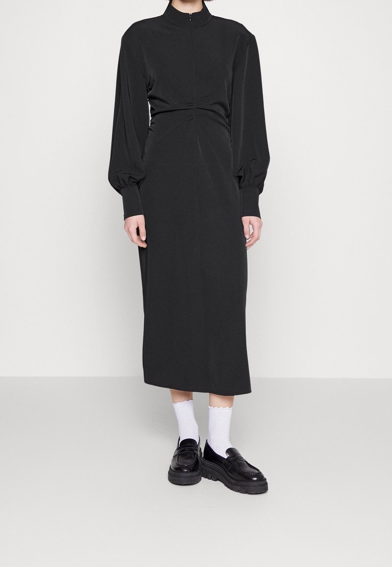 Femme portant une longue robe noire avec des manches longues bouffantes, des socquettes blanches à bords froufroutants et des mocassins plateforme noirs.