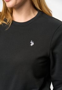 Svart sweatshirt i en bomullsblandning med rund halsringning och en litet vitt broderat logotyp på vänster bröstområde. Mjuk textur.