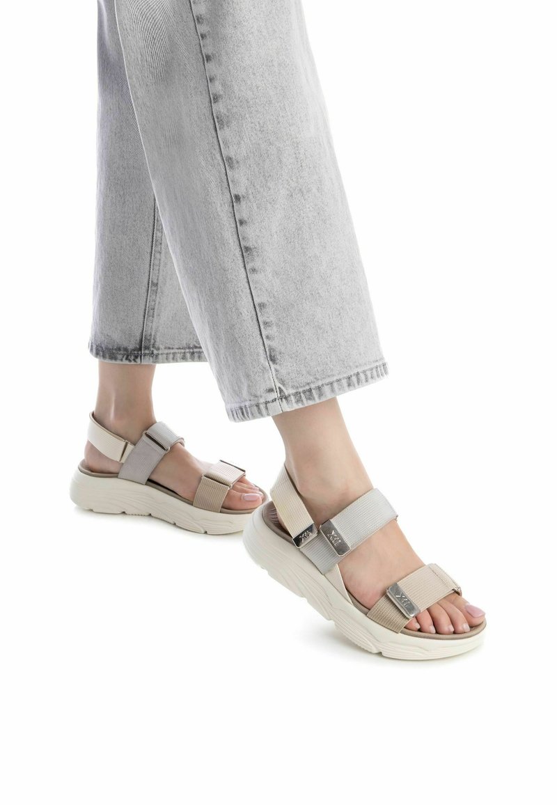 Sandalias beige claro con tiras texturizadas y hebillas ajustables, con una suela blanca gruesa y diseño de punta abierta, llevadas con denim gris.