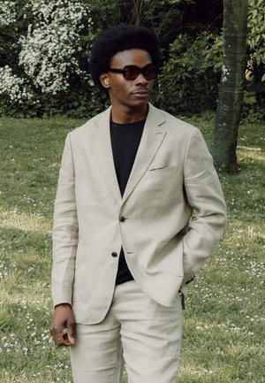 Mann mit beigem Anzug und schwarzem Hemd mit Sonnenbrille steht auf Gras nahe einem Baum und weißen Blumen im Hintergrund.