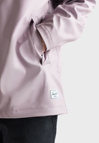 Giacca da pioggia rosa chiaro con una texture liscia, dotata di una tasca laterale e di un'etichetta bianca Herschel sull'orlo. Sono visibili pantaloni neri.
