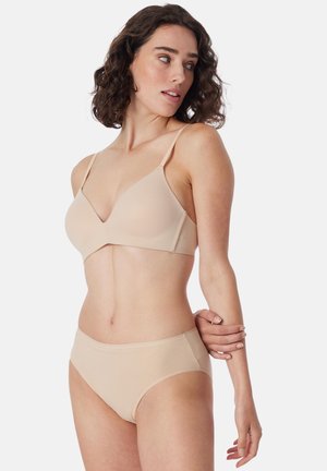 Woman with curly dark hair wearing nude seamless bra and matching underwear, looking sideways, holding her opposite arm.