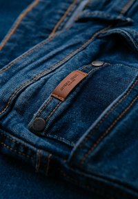 Vaqueros de mezclilla en azul oscuro con costuras naranjas. Presentan una etiqueta de marca de cuero y un detalle de botón metálico en la cintura.