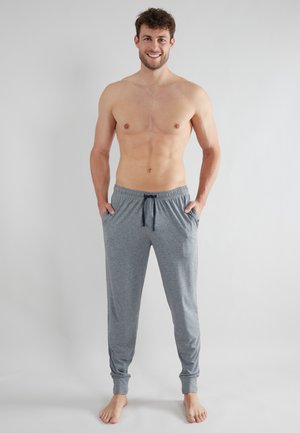 Oberkörperfreier Mann lächelnd, barfuß stehend mit Händen in den Taschen, trägt graue Loungebuxe mit Kordelzug vor einem einfachen, hellen Hintergrund.