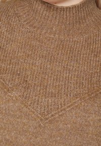 Pull marron en tricot avec un col montant côtelé et un motif texturé en forme de V sur la poitrine. Le tissu a une apparence douce et chaude.