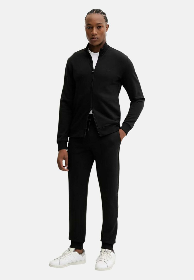 Svart zip-up jacka med ståkrage, i kombination med matchande svarta joggers och vita sneakers. Slitstark tyg, figurnära design, minimal detaljering.