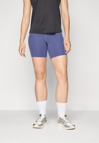 Shorts de sport violets au design ajusté, avec des coutures sur les côtés, associés à un haut sans manches noir et des baskets gris et blanches avec des chaussettes blanches.