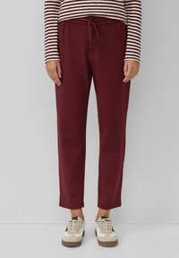 Persoon in bordeauxrode joggingsbroek, beige en witte sneakers, en een lange mouwen shirt met beige en bordeauxrode strepen, staande voor een effen achtergrond.