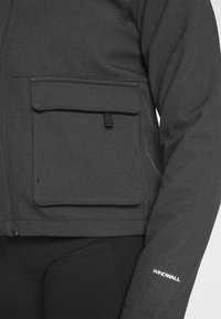 Grå dragkedjejacka i texturerat tyg med en ficka med klaff på framsidan och "WINDWALL"-logotyp på ärmen i vitt.