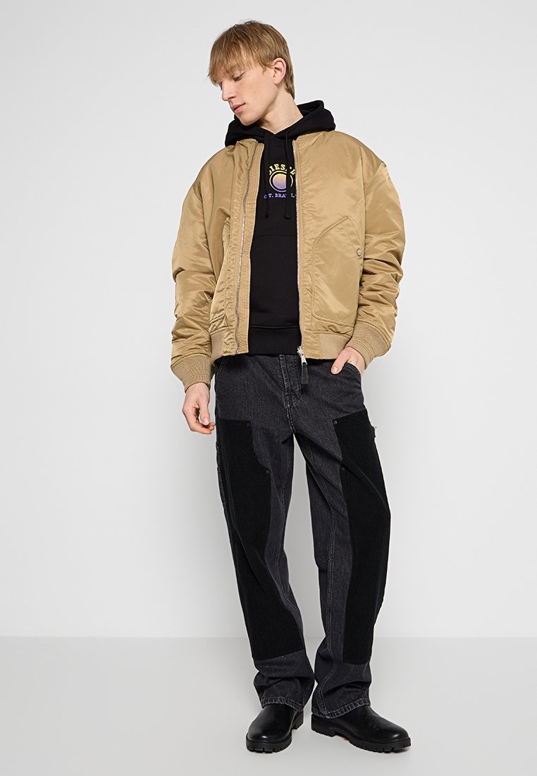 Giacca bomber beige in nylon, abbinata a una felpa nera con logo. Pantaloni in denim scuro con inserti a contrasto. Scarpe nere.