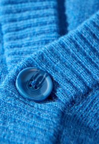Blau gestrickter Stoff mit gerippter Textur, versehen mit einem großen glänzenden blauen Knopf mit vier Löchern, der nahe dem Rand platziert ist.