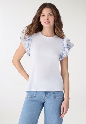 Donna con lunghi capelli ondulati che indossa una maglietta bianca con maniche a volant floreali blu e jeans azzurri, posando con una mano sul fianco.