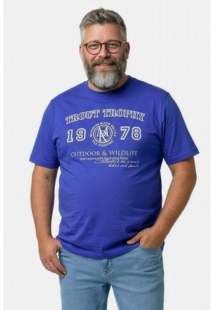 Lächelnder Mann mittleren Alters mit grauem Bart und Brille, trägt ein blaues T-Shirt mit der Aufschrift "Trout Trophy 1978" und hellblaue Jeans, die Hand in der Tasche.