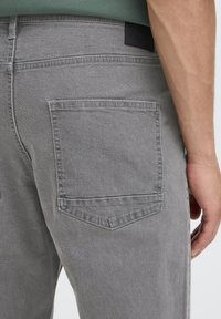 I pantaloni in denim grigio presentano una tasca applicata sul retro con forma rettangolare e cuciture rinforzate. Il tessuto sembra morbido con una trama opaca.
