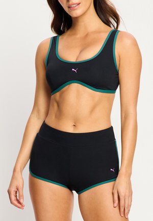 Femme portant un ensemble de brassière de sport noire et short avec bordure turquoise et logos Puma roses, debout devant un fond clair uni.
