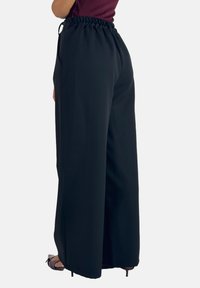 Zwarte wijde broek met een gatherings aan de elastische tailleband, van achteren weergegeven. Gladde stof met een losse pasvorm en geen zichtbare patronen.