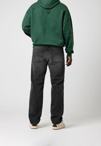 Man som bär en grön hoodie, svarta jeans och vita sneakers står vänd bort från kameran mot en enfärgad ljus bakgrund.