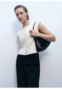 Cremfarbener ärmelloser Oberteil aus strukturiertem Stoff mit vorderen Knöpfen, kombiniert mit einer schwarzen geflochtenen Handtasche. Schwarze Maßanzughosen runden den Look ab.