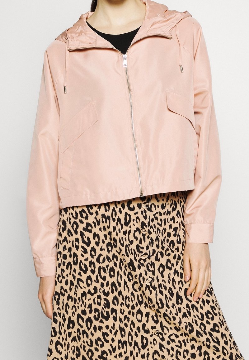 Personne portant une veste à capuche rose pâle zippée sur un haut noir, associée à une jupe beige et noire imprimée léopard à boutons.
