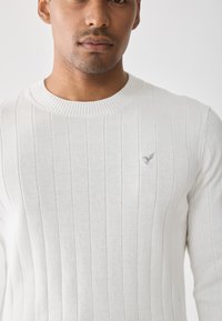 Pull en maille blanche avec un col rond. Comprend un tissu texturé et un petit emblème gris sur la poitrine. Design simple et épuré.