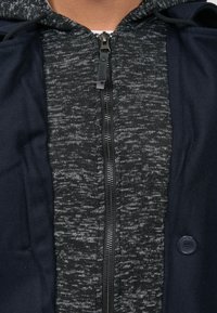 Marineblauwe capuchonjas met een textuur grijze gebreide binnenkant, ritssluiting en opvallend knoopdetail aan de rechterkant.