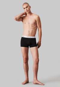 Schwarze Boxershorts mit einem weißen, markierten Bund, aus weicher Baumwolle, figurbetontem Schnitt und in mittlerer Oberschenkel-Länge. Das Modell posiert mit einer Hand am Hals.