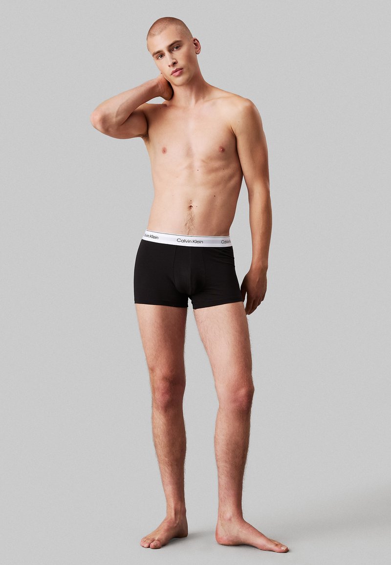 Schwarze Boxershorts mit einem weißen, markierten Bund, aus weicher Baumwolle, figurbetontem Schnitt und in mittlerer Oberschenkel-Länge. Das Modell posiert mit einer Hand am Hals.