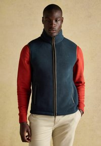 Tumši tirkīza krāsas fleeces veste ar augstu apkakli, rāvējslēdzēja aizdari un sānu kabatām, kombinēta ar sarkanu garām piedurknēm kreklu un gaišām biksēm.