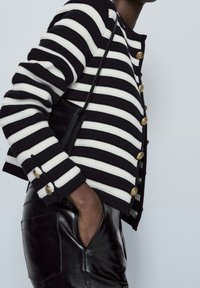 Cardigan a righe bianche e nere con texture a coste, bottoni dorati e pantaloni in pelle nera, con dettagli su tasca laterale.