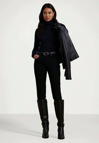 Fekete garbót, fekete övvel ellátott nadrágot és térd fölé érő fekete csizmát viselő nő, akinek egy fekete bőrkabát van az egyik vállán.