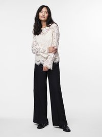 Ivoor kanten blouse met lange mouwen, een geschulpte zoom en ingewikkelde bloemenpatronen, gecombineerd met wijde zwarte broek en zwarte schoenen.