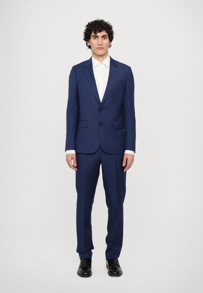 Un costume bleu marine avec un revers cranté, fermeture à un bouton, et deux poches avant, associé à une chemise blanche et des chaussures noires.