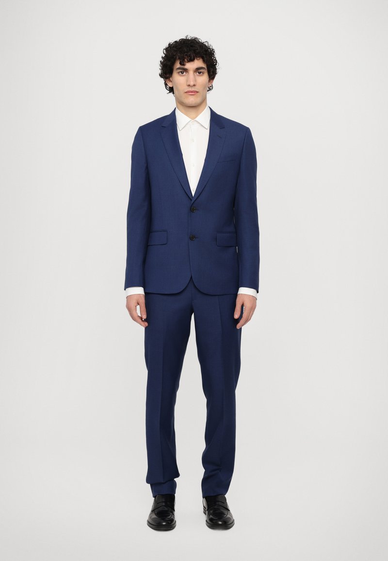 Un costume bleu marine avec un revers cranté, fermeture à un bouton, et deux poches avant, associé à une chemise blanche et des chaussures noires.
