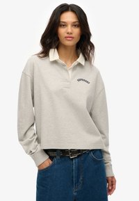 Polo corto de color gris claro con un cuello blanco y el logo bordado de "Superdry". Cuenta con mangas largas y un corte relajado. Jeans de mezclilla azul visibles.