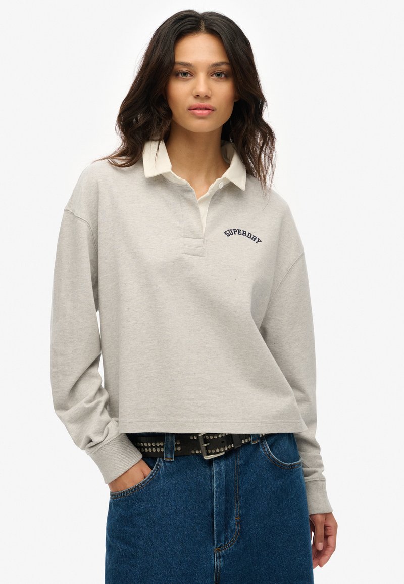 Polo corto de color gris claro con un cuello blanco y el logo bordado de "Superdry". Cuenta con mangas largas y un corte relajado. Jeans de mezclilla azul visibles.