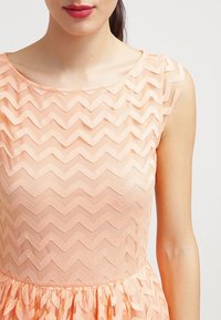 Robe couleur pêche avec un motif en zigzag, haut transparent et jupe en tissu superposé. Texture douce et design de corsage ajusté.