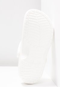 Sabots blancs en caoutchouc avec une semelle texturée, présentant un design traditionnel et une sangle au dos. L'empeigne arbore des courbes douces et des trous de ventilation.