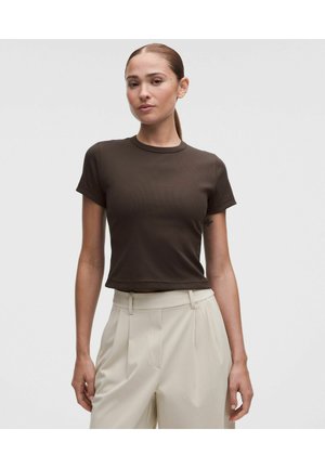 Mujer con el cabello recogido, vistiendo una camiseta ajustada de manga corta color marrón oscuro y pantalones plisados de cintura alta color beige claro sobre fondo gris.